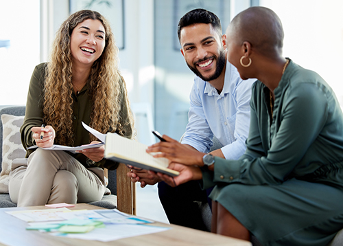 Three happy business colleagues of diverse ethnicity holding paperwork and smiling at each other during a meeting around a low (coffee) table.