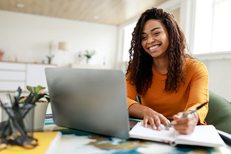 Happy young woman at laptop watching webinar and taking notes.